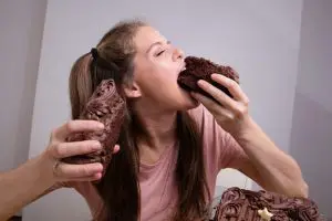 Woman eating a big piece of chocolate cake from one hand with another piece in opposite hand, representing pleasure principle